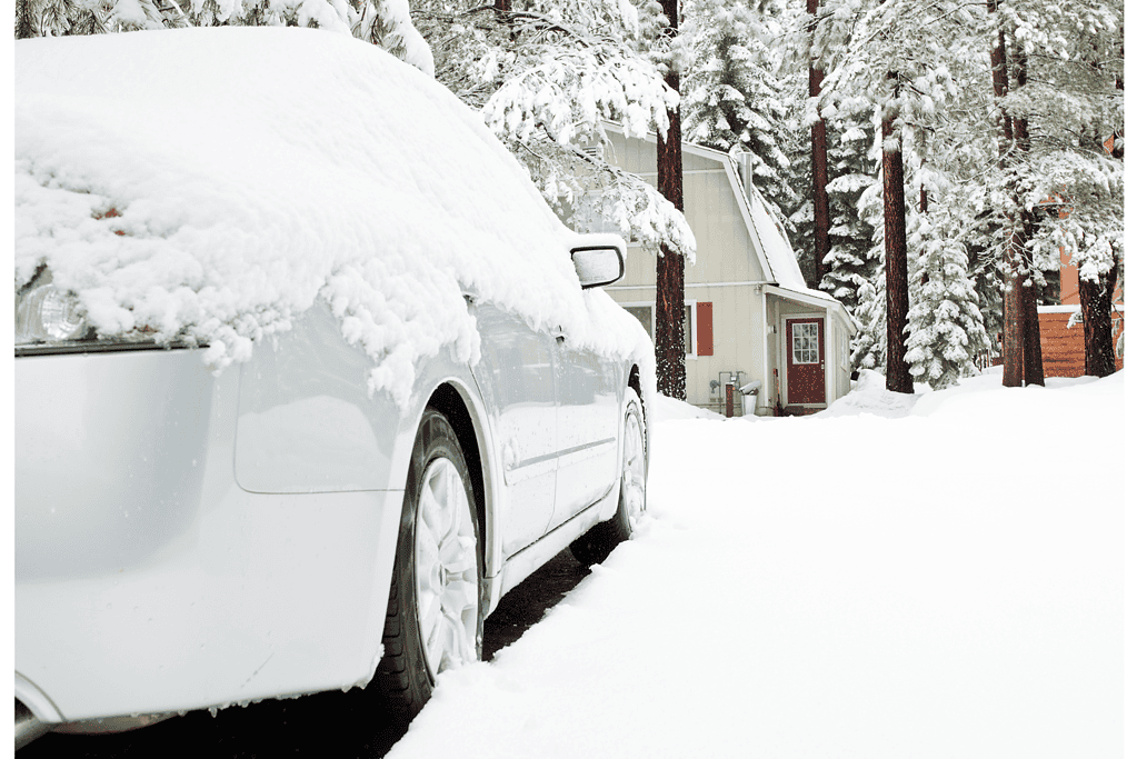 White car covered in snow in a winter landscape with trees and a house.